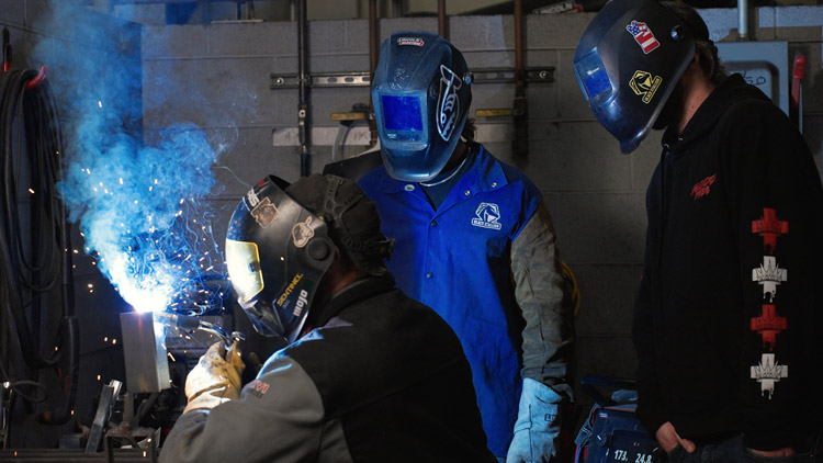 Two students watch a teacher weld.